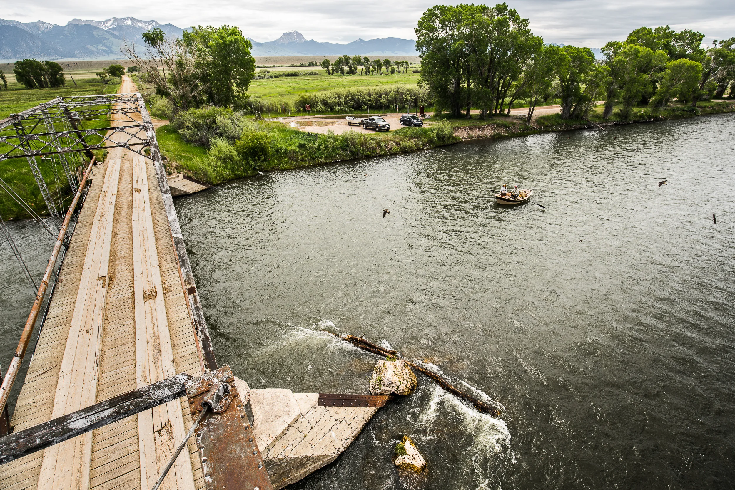 Drift boat passing under a wooden bridge with mountain views